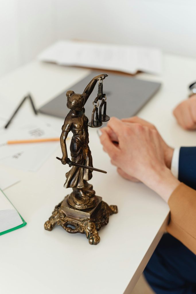 pexels-photo-7876148-7876148 Bronze Lady Justice statue on a white office desk with hands in the background. Symbol of law and fairness.